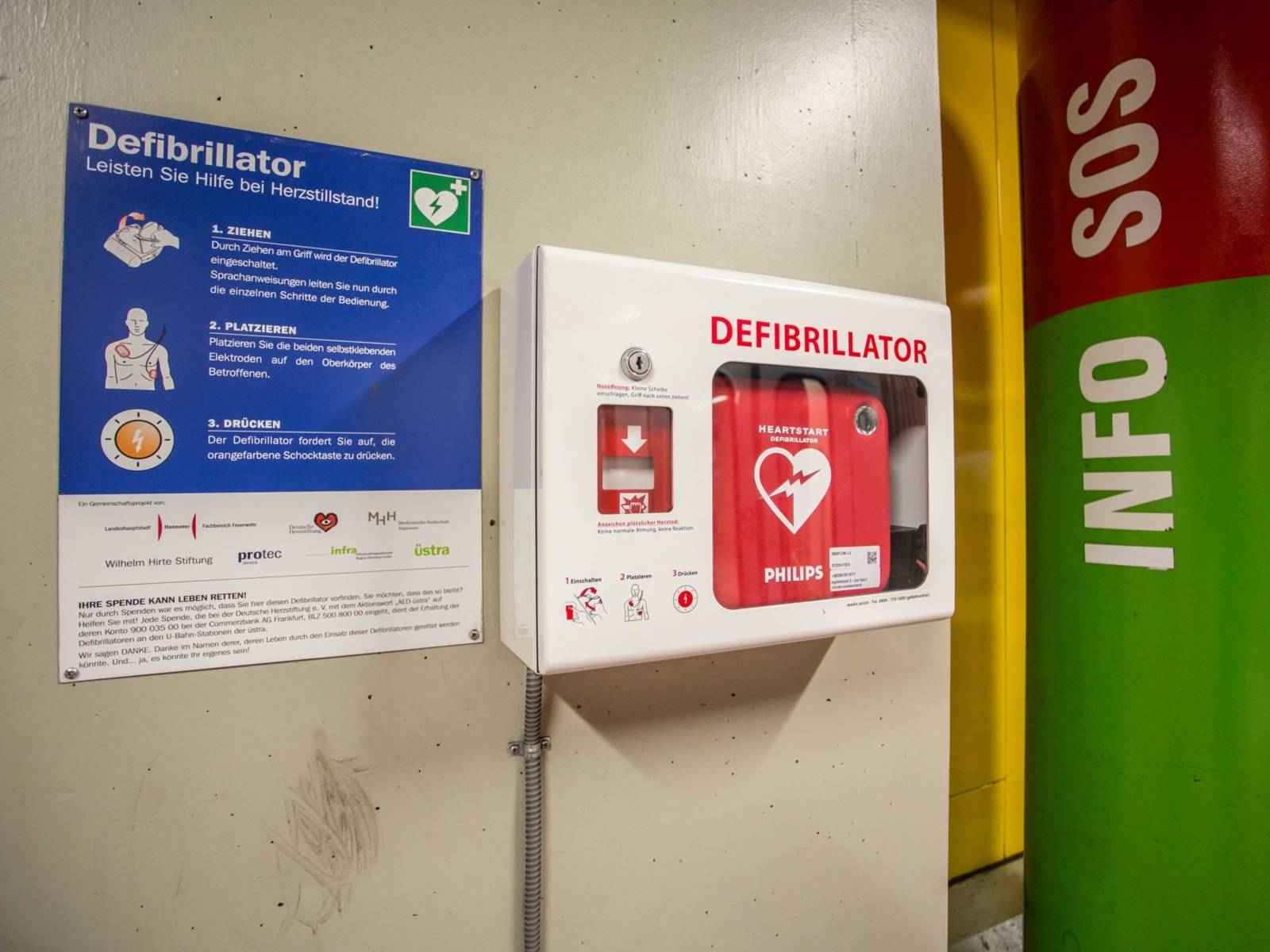 Ein Defibrillator an der Wand, daneben eine Notrufsäule