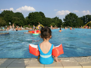 Mädchen mit Schwimmflügeln sitzt mit dem Rücken zum Betrachter am Beckenrand. Im Freibad plantschen viele Kinder.