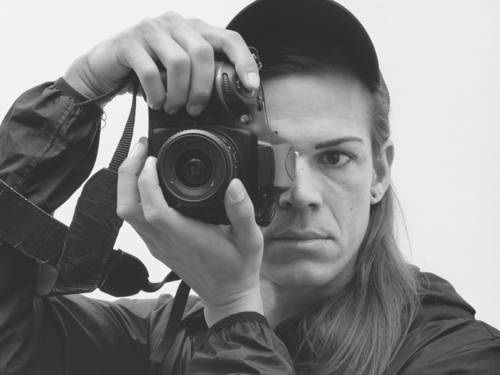 Zu sehen ein schwarz-weißes Foto einer Person mit einer Kamera im Gesicht, die sich selbst fotografiert.