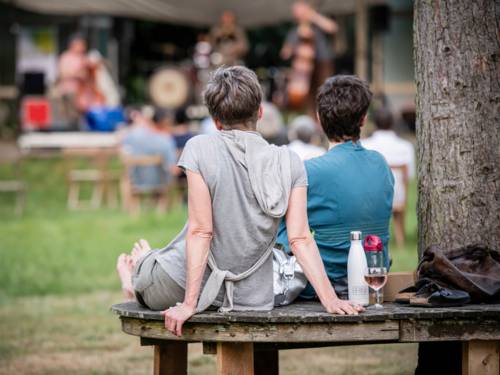 Zwei Personen sitzen entspannt draußen an einem Baum auf einer Bank und lauschen einem Konzert
