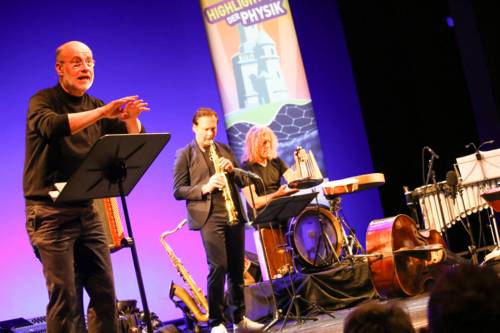 Harald Lesch am Rednerpult, im Hintergrund zwei Musiker des Quartetts.