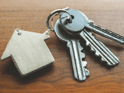 Drei Schlüssel an einem Schlüsselring liegen auf einer Holzplatte, daran ein aus Holz gesägter Anhänger in der Form eines Hauses.