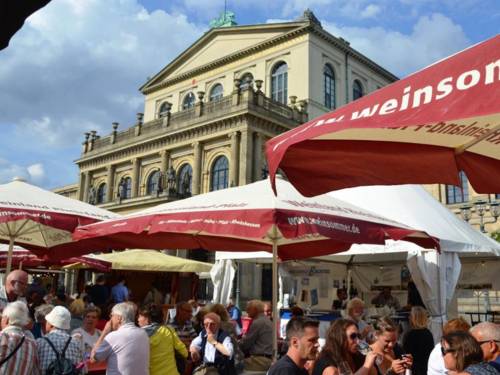 Menschen sitzen auf Banken unter Sonnenschirmen auf einem Platz vor einem historischem  Gebäude und trinken aus Weingläsern.