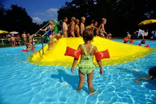Das Kinderplantschbecken, im Vordergrund ein kleines Mädchen mit hellgrünem Badeanzug und roten Schwimmflügel, dahinter eine niedrige, kleine gelbe Rutsche