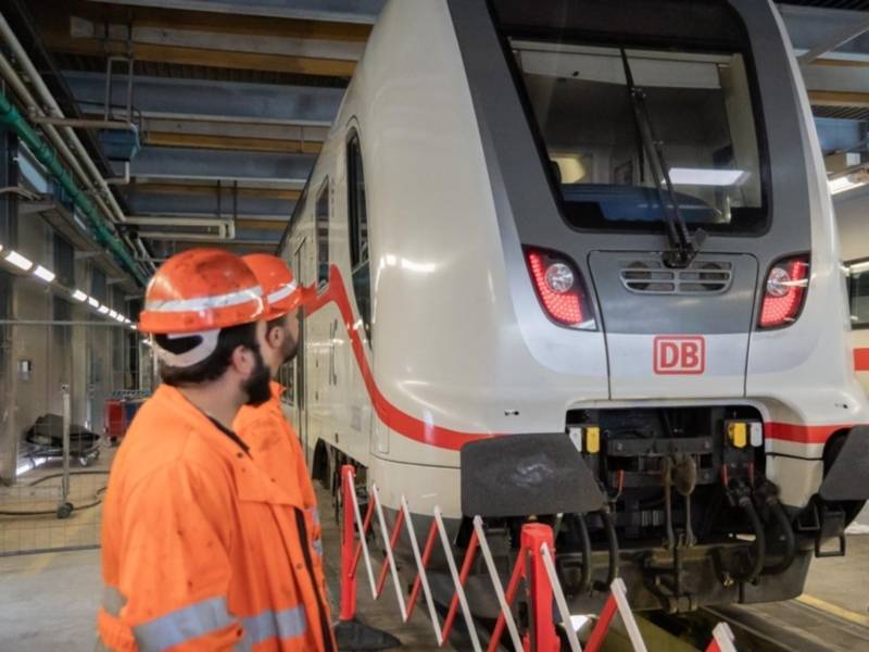 Zwei Männer in oranger Sicherheitskleidung mit Helm sehen in einer Halle auf einen DB-Zug.