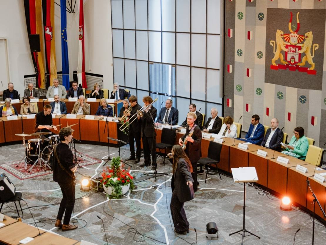 Eine Band spielt in der Mitte eines Plenarsaals. Die MusikerInnen stehen im Kreis zueinander. In der rechten oberen Bildecke ein Wandteppich mit dem Wappen der Landeshauptstadt Hannover.
