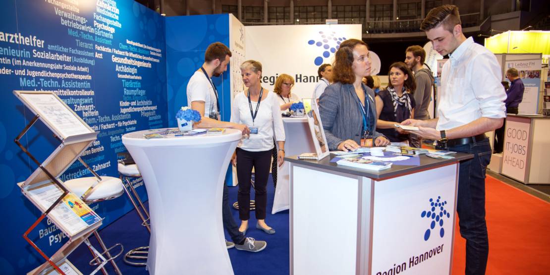 Ein Mann informiert sich an einem Messestand. Infomaterial liegt aus und Standpersonal beantwortet Fragen.