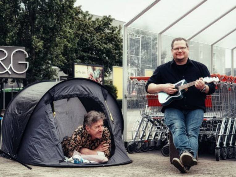 Ein Mann mit Gitarre sitzt vor einem Plastikgehäuse mit ineinandergeschobenen Einkaufswagen, neben ihm liegt ein zweiter bäuchlings in einem kleinen Zelt.