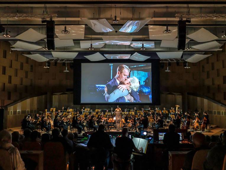 Filmszene mit sich küssendem Paar auf einer Leinwand über einem Orchester.