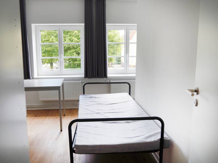 Vorn eine offene Tür, dahinter ein Bettgestell mit einer Matratze, links im Anschnitt ein Spind. Links steht ein Tisch, geradeaus zwei Fenster.
