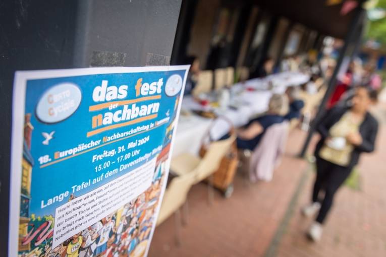 Im Vordergrund des Bildes ist ein Plakat zu sehen. Die Aufschrift ist: "Das Fest der Nachbarn - 18. Europäischer Nachbarschaftstag in Hannover". Im Hintergrund des Bildes sind, unscharf, mehrere Menschen zu sehen.