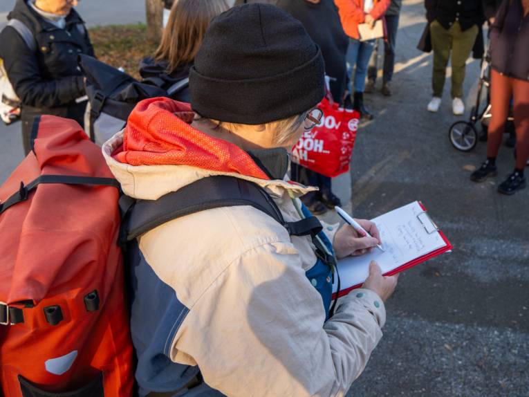 Eine Frau mit Rucksack und Mütze steht auf einem Fußweg und schreibt auf ein Papier auf einem Klemmbrett. Um sie stehen weitere Personen.