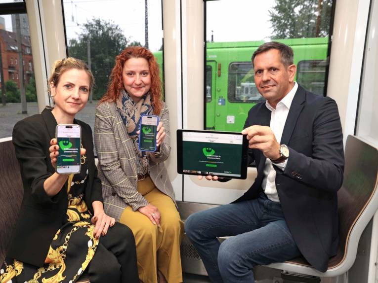 Zwei Frauen und ein Mann sitzen in einer Stadtbahn und halten mobile Endgeräte in die Kamera.