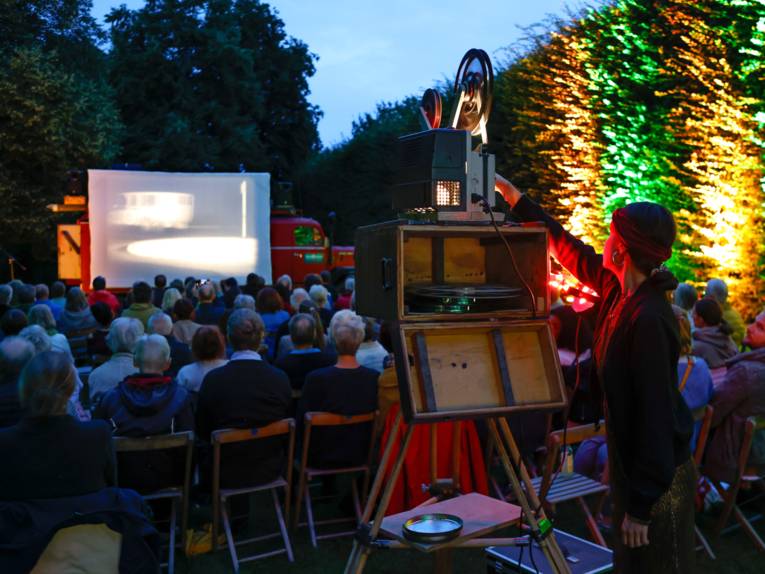 Ein Projektor wirft die Bilder eines Films auf eine Leinwand bei einer Veranstaltung unter freiem Himmel, Publikum schaut den Film. Eine Frau legt Hand an die Technik des Projektors.