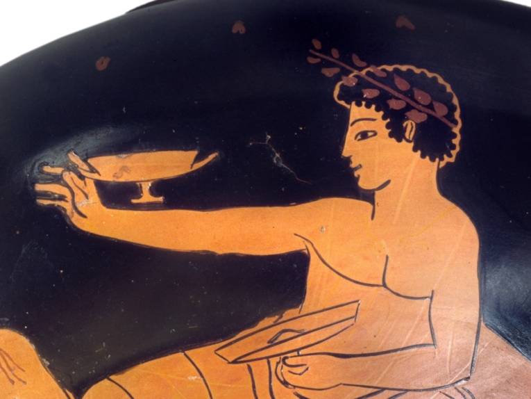Griechischer Aristokrat in seitlicher Lage und Profilansicht beim Symposion mit jeweils einer Trinkschale in der Hand. Der rechte Arm ist seitlich ausgestreckt.