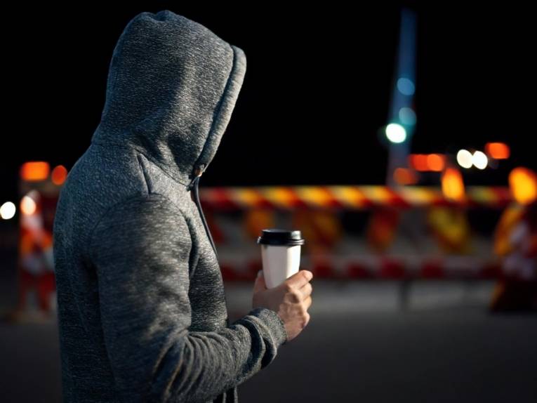 Ein Mann von der Seite mit Kapuze steht im Dunkeln auf einer Straße und hält eine Kaffeetasse. Dahinter sind Baustellenabsperrungen zu sehen.