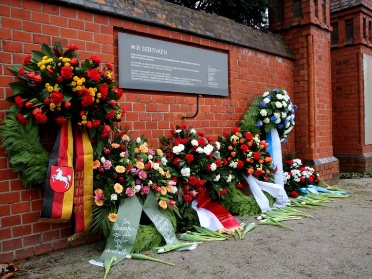 Sechs Blumenkränze abgelegt vor einer Gedenkwand.