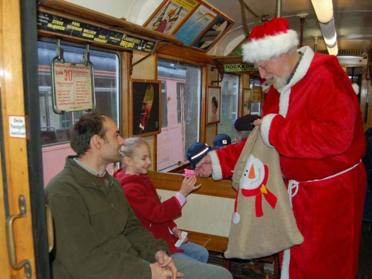 Mann und Mädchen sitzen in einer historischen Straßenbahn, ein Mann in einem Nikolauskostüm gibt dem Mädchen ein Geschenk.